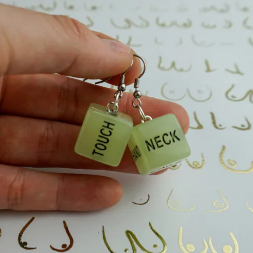 Someone holds a pair of our glow in the dark sex dice earrings. They say the words "touch" and "neck". The background is white, with a repeated pattern of boobs drawn in gold.