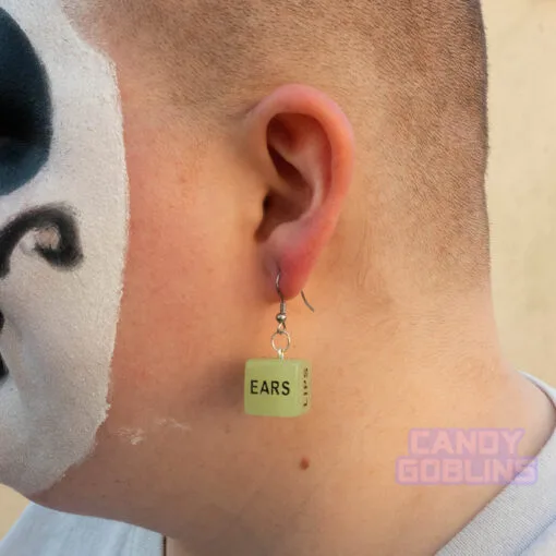A side view of a model wearing black and white juggalo clown makeup. In their ear they are wearing a sex dice earring, and the word "ears" can be seen.