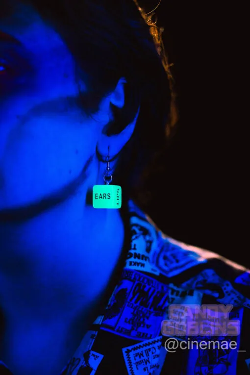 A model under a blue blacklight, they are wearing our sex dice earrings and they are glowing. The word "ears" and "lips" can be seen on the surface of the die.