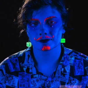 A model under a blue blacklight, they have a neon orange moustache, beard, and eyebrows painted on. They are wearing our sex dice earrings and they are glowing bright green. The word 