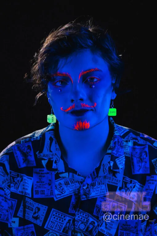 A model under a blue blacklight, they have a neon orange moustache, beard, and eyebrows painted on. They are wearing our sex dice earrings and they are glowing bright green. The word "ears" and "suck" can be seen on the surface of the die.