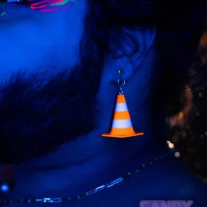 A black light photo of a person's ear, with a traffic cone earring dangling from it. The orange in the traffic cone has gone fluorescent under the black light, making it stand out.