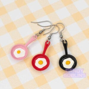 Our egg pan earrings are displayed together. The pans are red, black, and pink, and all have an egg frying on the surface.