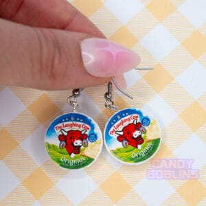 Someone holds a pair of Laughing Cow earrings, they have a decorative pink nail