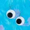 A pair of googly eye earrings are laid against a blue furry background. The earrings are black and white.