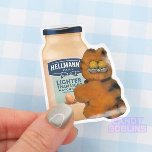 A hand holds a sticker of a bizarre looking Garfield toy holding on a jar of mayonnaise.