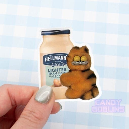 A hand holds a sticker of a bizarre looking Garfield toy holding on a jar of mayonnaise.