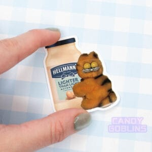 A hand holds a sticker of a bizarre looking Garfield toy holding on a jar of mayonnaise.