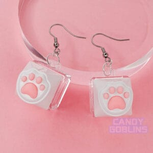 A pair of our Cat Keyboard Switch Earrings posed on a glass photo prop. The earrings are leaning so you can see the titanium hooks clearly. The background is a pastel pink.