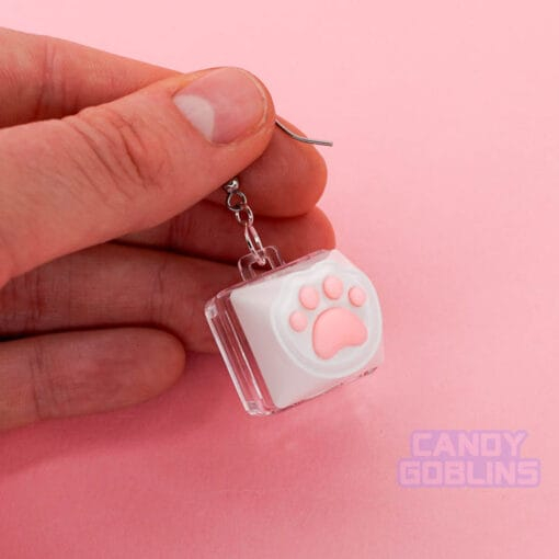 A hand holds a single Cat Keyboard Switch Earring. The background is a pastel pink.