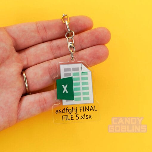 A hand holds our Excel Keychain. The background is yellow. The keychain features the Microsoft Excel logo, with text underneath that reads "asdfghj FINAL FILE 5.xlsx"