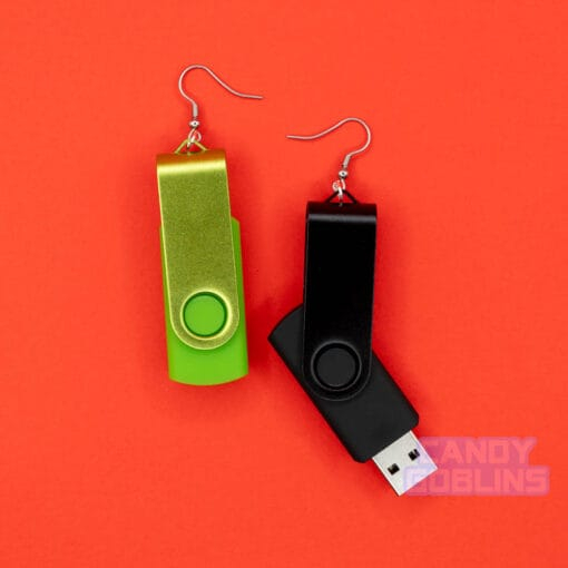 A pair of USB stick earrings, one USB is black, the other is a bright green. The green USB stick has been folded away, while the black one is unfolded. They are photographed against a red background.