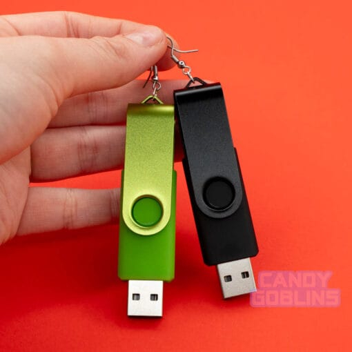 A hand holding a pair of USB stick earrings, one USB is black, the other is a bright green. They are photographed against a red background.