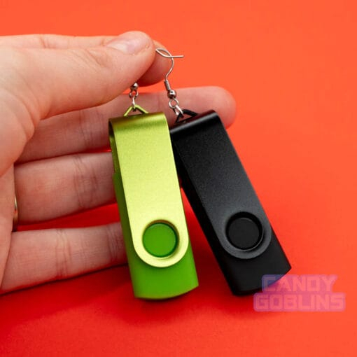 A hand holding a pair of USB stick earrings, one USB is black, the other is a bright green. They are photographed against a red background.