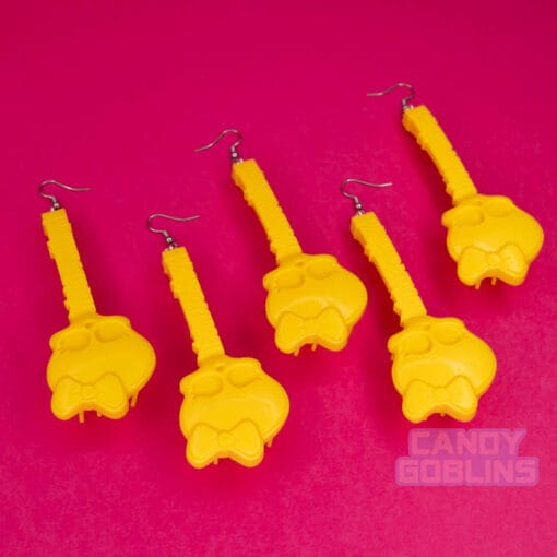 Five yellow doll brush earrings are laid out against a pink background. The handles say "monster high" and feature a skull wearing a bow on it's head.