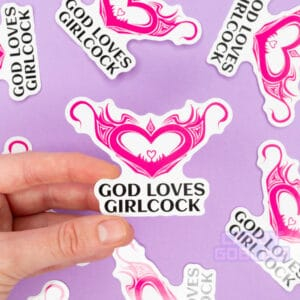 A person holds a sticker that says god loves girlcock and features a hentai womb tattoo. The background is lilac and you can see other version of the same sticker scattered around.
