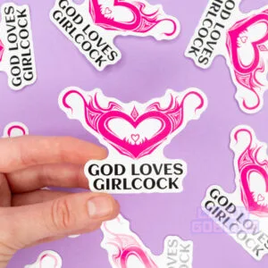 A person holds a sticker that says god loves girlcock and features a hentai womb tattoo. The background is lilac and you can see other version of the same sticker scattered around.