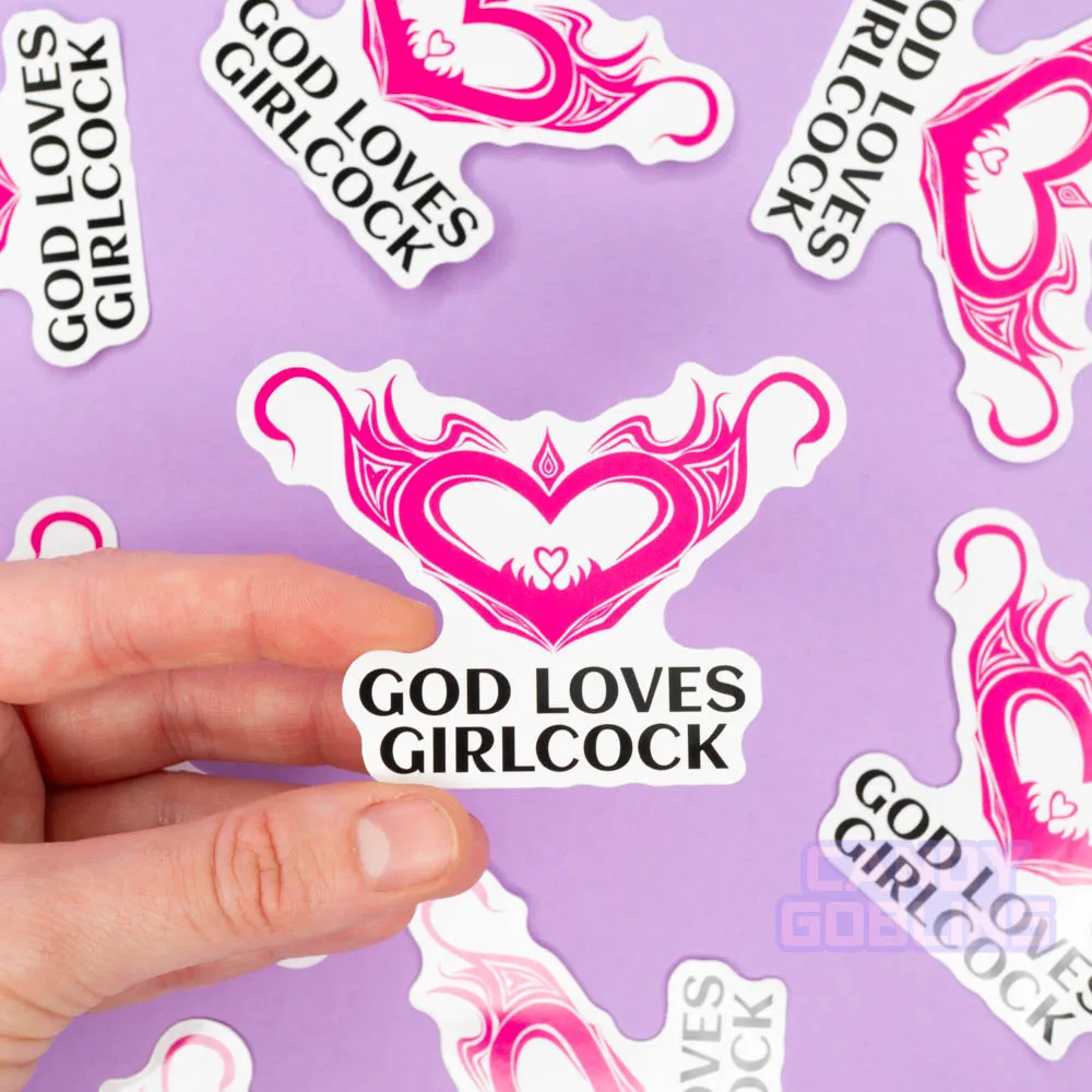 A person holds a sticker that says god loves girlcock and features a hentai womb tattoo. The background is lilac and you can see other version of the same sticker scattered around.