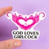 A person holds a sticker of a hot pink womb heart design. The text underneath reads "god loves girlcock"