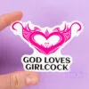 A person holds a sticker of a hot pink womb heart design. The text underneath reads "god loves girlcock"
