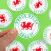 A person holds up a circle sticker with a Welsh dragon in the middle, text around it reads "Proud Welsh Faggot"