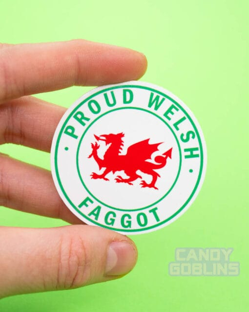 A person holds up a circle sticker with a Welsh dragon in the middle, text around it reads "Proud Welsh Faggot"