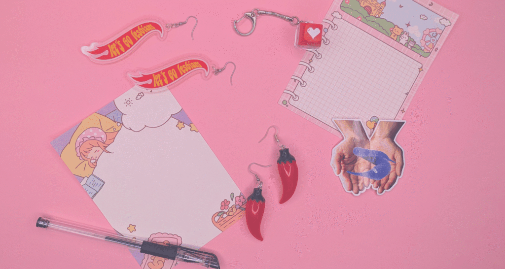 Note paper, pens, earrings, and keychains are scattered against a pink backdrop.