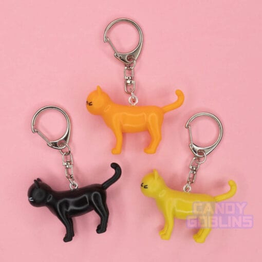 Cat keychains lay against a pink backdrop. The cats are orange, yellow, and black