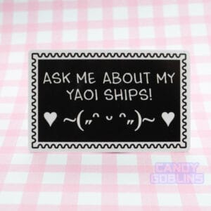 A stamp shaped sticker reads: Ask Me About My Yaoi Ships. Under the text is a happy emoticon, blushing and dancing with hearts. It's against a pink background.