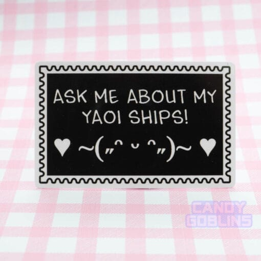 A stamp shaped sticker reads: Ask Me About My Yaoi Ships. Under the text is a happy emoticon, blushing and dancing with hearts. It's against a pink background.
