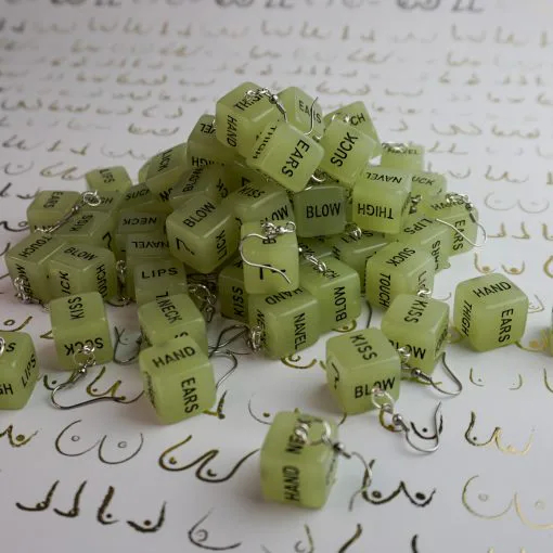 A massive pile of our glow in the dark sex dice earrings. There's probably at least 10 pairs stacked up. The background is white, with a repeated pattern of boobs drawn in gold.