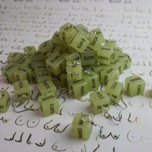 A massive pile of our glow in the dark sex dice earrings. There's probably at least 10 pairs stacked up. The background is white, with a repeated pattern of boobs drawn in gold.