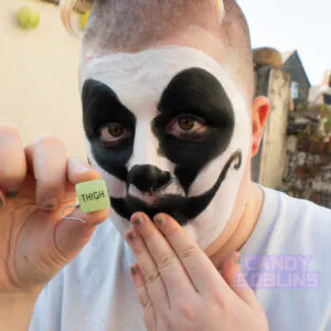 A model with black and white juggalo clown makeup holds up a sex dice earring. The word 