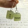 Someone holds a pair of our glow in the dark sex dice earrings. They say the words "lick" and "thigh". The background is white, with a repeated pattern of boobs drawn in gold.
