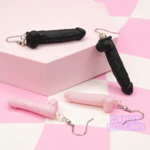 A pair of black dildo earrings and pink dildo earrings are arranged against a pink photo prop.