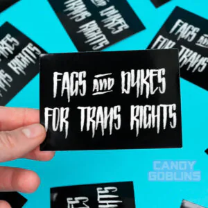 A Fags and Dykes for Trans Rights sticker is held up by a lesbian. In the background you can see more of the same sticker.