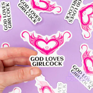 A person holds a sticker that says god loves girlcock and features a hentai womb tattoo. The background is lilac and you can see other version of the same sticker scattered around.