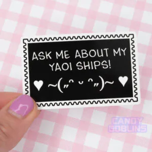 Someone holds a sticker shaped like a deviantart stamp that reads Ask Me About My Yaoi Ships. Under the text is a happy emoticon, blushing and dancing with hearts.
